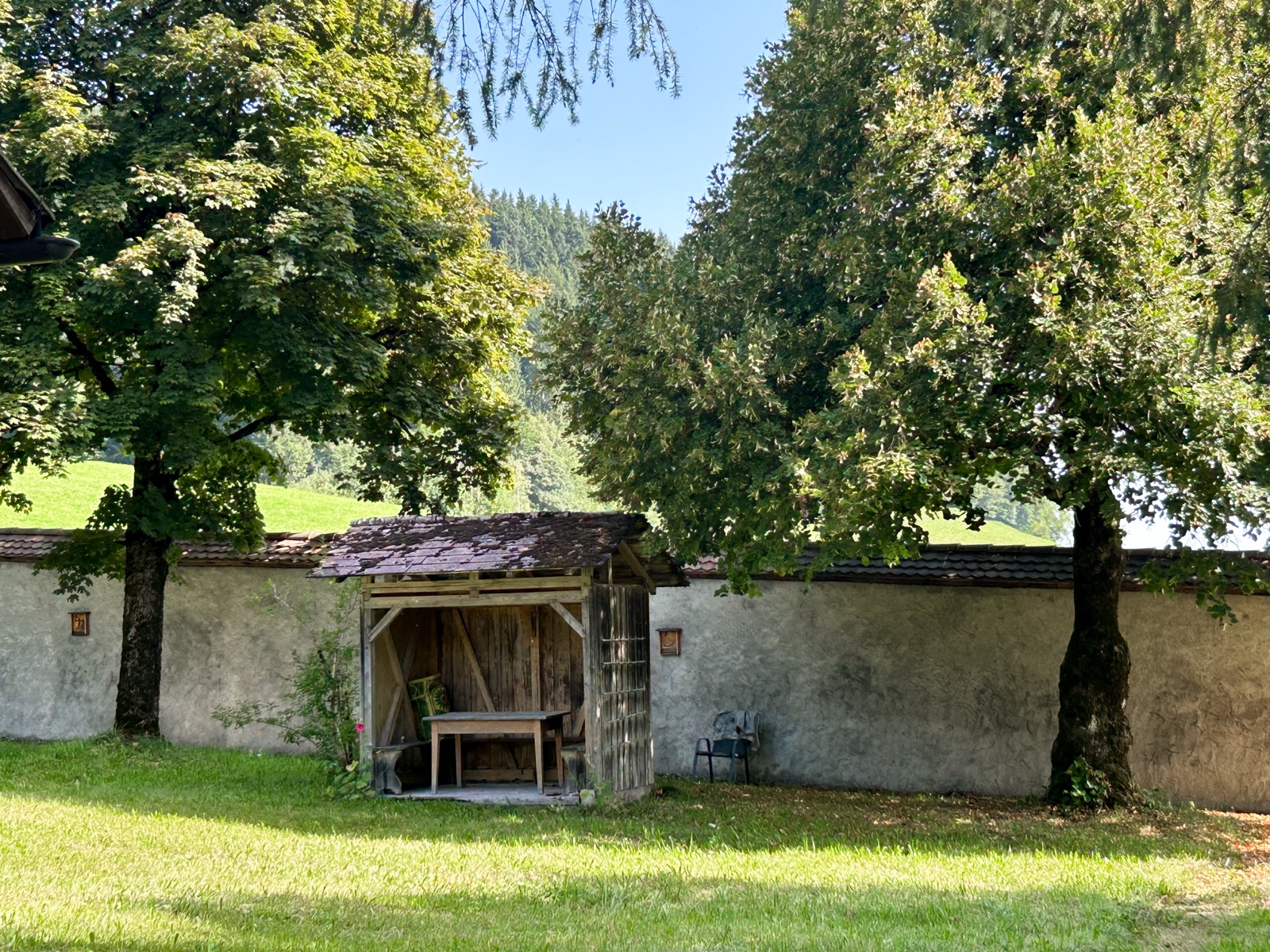 Holzlaube an der Klostermauer, umgeben von alten Bäumen und Wiese